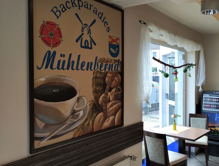 Mühlenbernd Valdorf Innenraum.jpg Innenansicht einer Bäckerei mit einem Tisch, Kaffeeautomat und einem dekorativen Wandbild mit Logo.