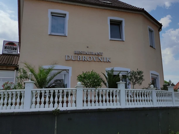 Außenansicht Dubrovnik Restaurant Dubrovnik mit beigefarbener Fassade, weißem Zaun und umliegender Pflanzenpracht.