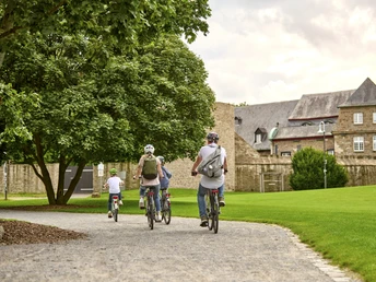 MüGa / Darlington-Park, Mülheim an der Ruhr Vier Radfahrende auf einem Weg im Park, flankiert von Bäumen und historischem Gebäude im Hintergrund.