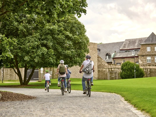 MüGa / Darlington-Park, Mülheim an der Ruhr Vier Radfahrende auf einem Weg im Park, flankiert von Bäumen und historischem Gebäude im Hintergrund.