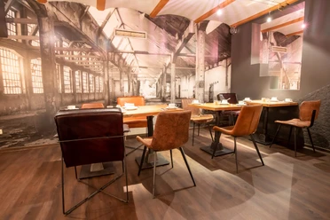Möbelei Restaurant mit Industriecharme: Verschiedene Stühle, Holztische, große Fototapete einer Fabrik.