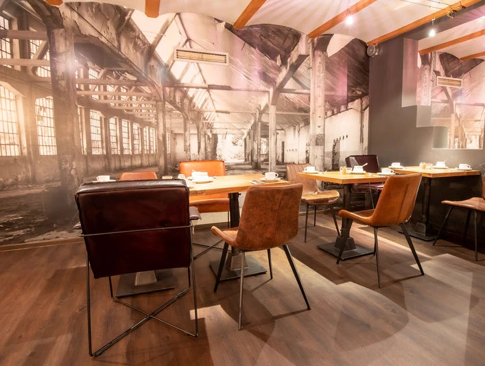 Möbelei Restaurant mit Industriecharme: Verschiedene Stühle, Holztische, große Fototapete einer Fabrik.