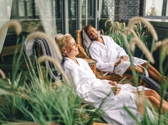 Paar in weißen Bademänteln entspannt auf Liegen auf einem begrünten Balkon.