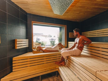 Ishara: Valeosauna Ein Mann und eine Frau entspannen in einer modernen Sauna mit hölzernem Interieur und großem Fenster.
