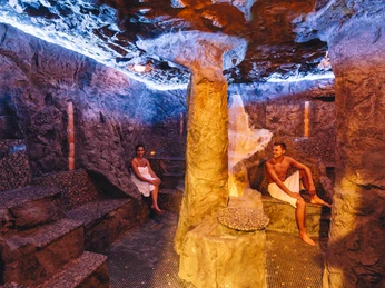 Ishara: Hoehlensauna Zwei Personen sitzen in einer stimmungsvoll beleuchteten Höhlensauna mit steinernen Wänden.