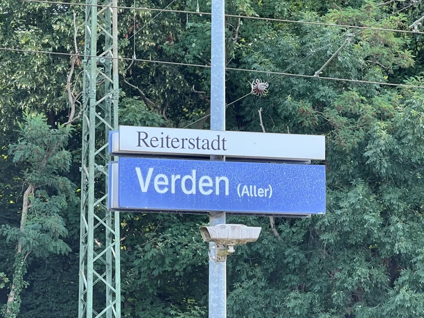 Bahnhofsschild mit der Aufschrift "Verden (Aller)" unter grünen, von Bäumen umgebenen Oberleitungen.