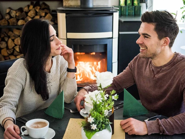 Gastraum Ottomühle Ein Paar sitzt lächelnd an einem Tisch mit Kaffee vor einem Kamin; im Vordergrund steht ein Blumenarrangement, die Atmosphäre ist gemütlich.