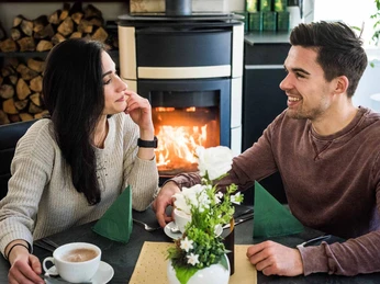 Ottomühle restaurant Ein Paar sitzt lächelnd an einem Tisch mit Kaffee vor einem Kamin; im Vordergrund steht ein Blumenarrangement, die Atmosphäre ist gemütlich.A couple sits smiling at a table with coffee in front of a fireplace; in the foreground is a flower arrangement, the atmosphere is cozy.Pár sedí s úsměvem u stolu s kávou před krbem, v popředí je květinové aranžmá, atmosféra je útulná.Para siedzi uśmiechnięta przy stoliku z kawą przed kominkiem; na pierwszym planie jest kompozycja kwiatowa, atmosfera jest przytulna.Een koppel zit glimlachend aan een tafel met koffie voor een open haard; op de voorgrond staat een bloemstuk, de sfeer is gezellig.Una coppia siede sorridente a un tavolo con un caffè davanti a un camino; in primo piano c'è una composizione floreale, l'atmosfera è accogliente.