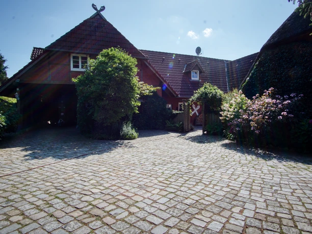 Gepflasterter Hof vor einem roten Backstein-Bauernhaus mit grünem Garten.