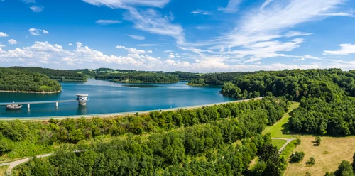 Große Dhünntalsperre Eine malerische Landschaft mit ruhigem See, umgeben von dicht bewaldeten Hügeln unter blauem Himmel.