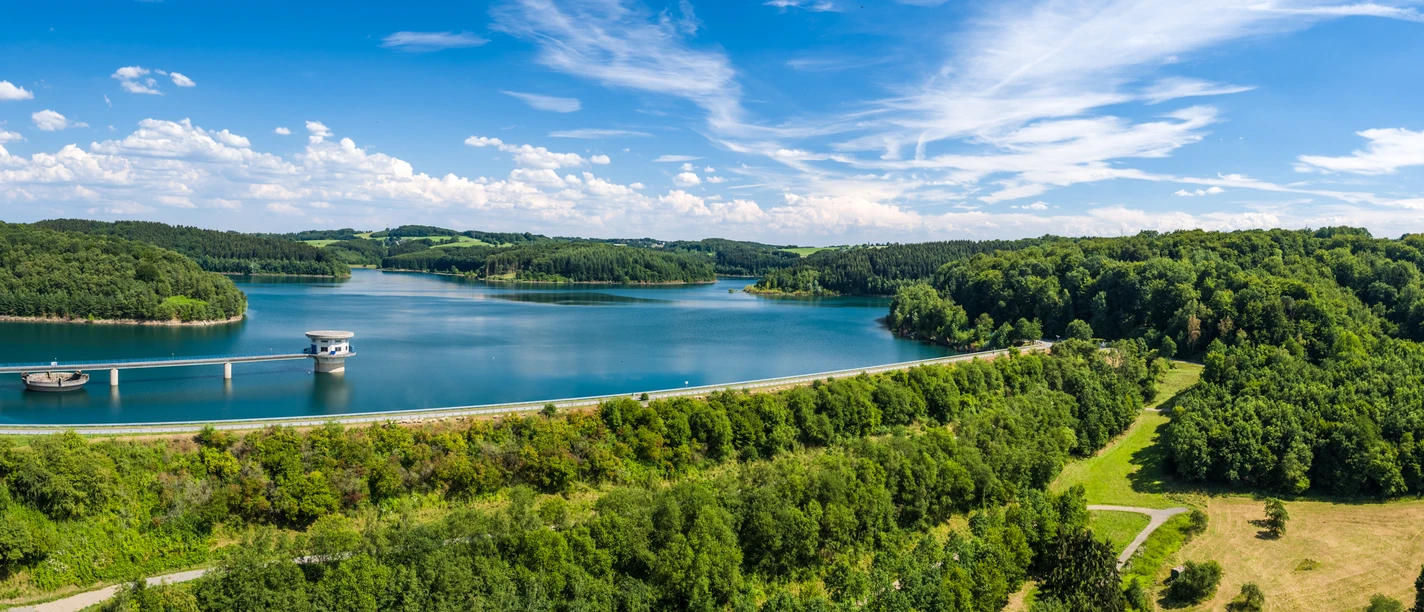 Große Dhünntalsperre Eine malerische Landschaft mit ruhigem See, umgeben von dicht bewaldeten Hügeln unter blauem Himmel.
