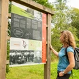 Infotafel am Böllweg Eine Frau betrachtet ein hölzernes Informationsschild in einem grünen Park mit Bäumen im Hintergrund.