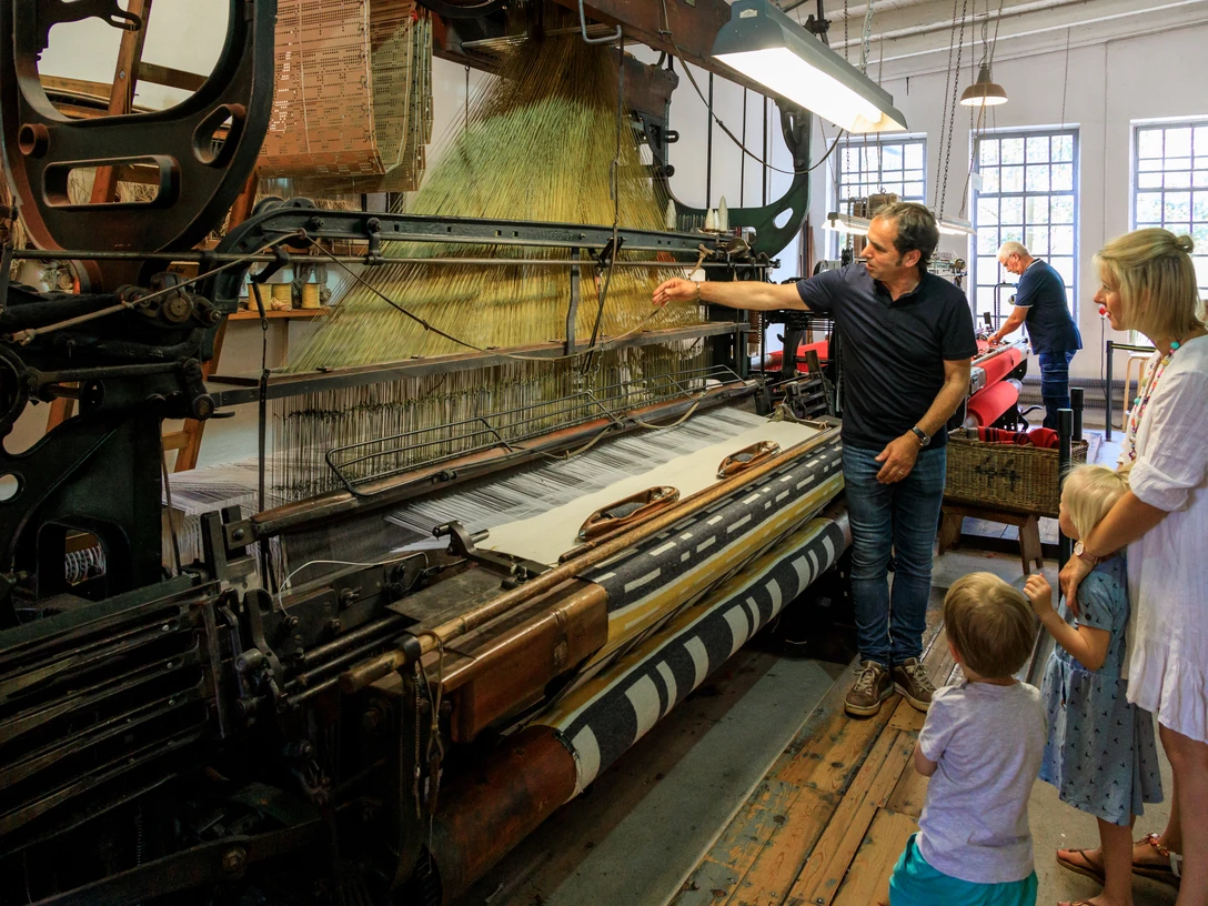 Führung an laufenden Maschinen Historische Webmaschine mit Besuchergruppe, darunter Erwachsene und Kinder, in einem Museum.Historical weaving machine with a group of visitors, including adults and children, in a museum.Historisk vævemaskine med en gruppe besøgende, herunder voksne og børn, på et museum.Historische weefmachine met een groep bezoekers, waaronder volwassenen en kinderen, in een museum.