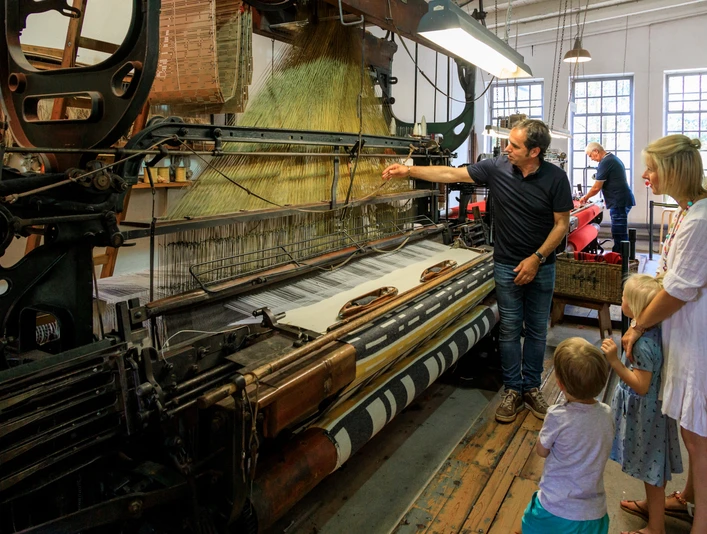Führung an laufenden Maschinen Historische Webmaschine mit Besuchergruppe, darunter Erwachsene und Kinder, in einem Museum.Historical weaving machine with a group of visitors, including adults and children, in a museum.Historisk vævemaskine med en gruppe besøgende, herunder voksne og børn, på et museum.Historische weefmachine met een groep bezoekers, waaronder volwassenen en kinderen, in een museum.