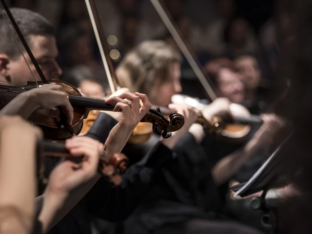 Nahaufnahme eines Sinfonieorchesters in Aktion, Geiger konzentriert beim Spielen, warme Beleuchtung.