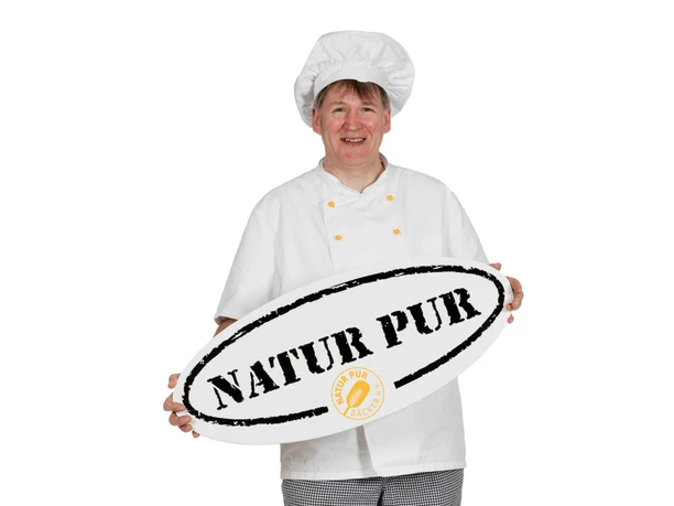Bäckermeister in Arbeitskleidung Bäckermeister in Arbeitskleidung hält lächelnd ein ovales Schild mit der Aufschrift „NATUR PUR“.
