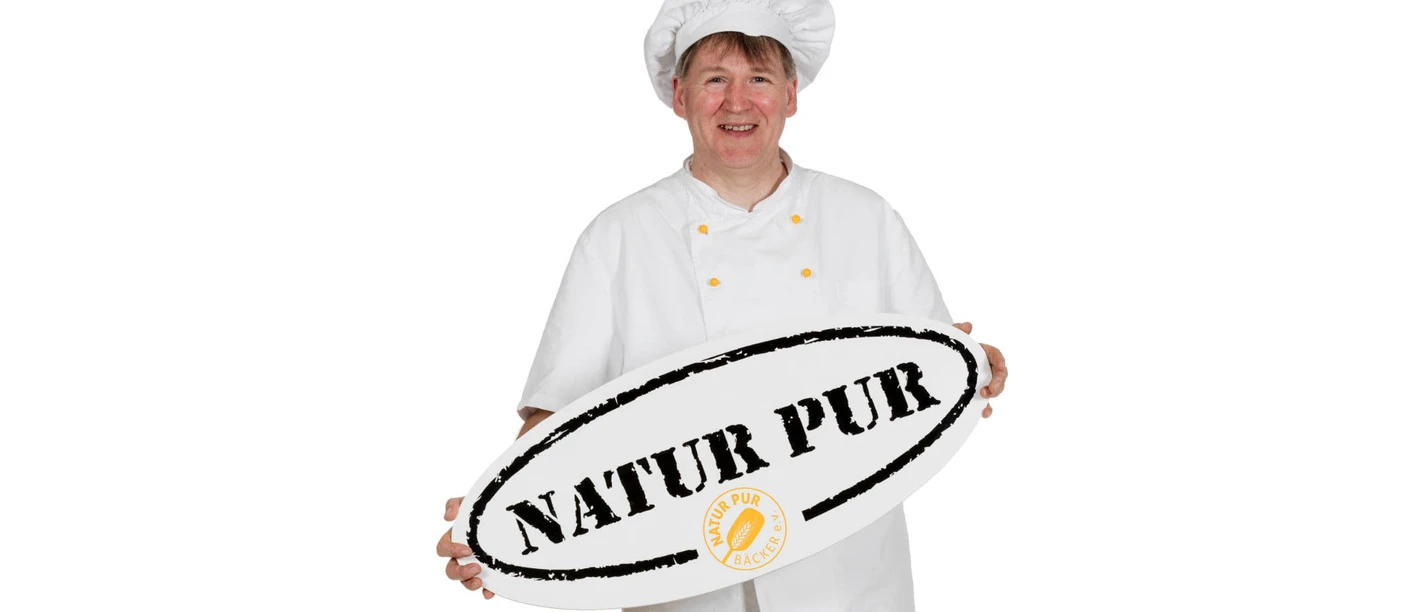 müller-bäcker-oldendorf-luhe.jpg Bäckermeister in Arbeitskleidung hält lächelnd ein ovales Schild mit der Aufschrift „NATUR PUR“.