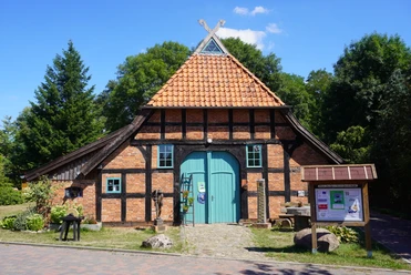 Backsteinfachwerkhaus mit dunklen Holzbalken und rotem Ziegeldach neben Infotafel und Bäumen.
