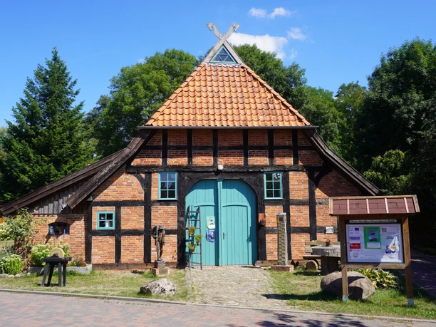 Backsteinfachwerkhaus mit dunklen Holzbalken und rotem Ziegeldach neben Infotafel und Bäumen.