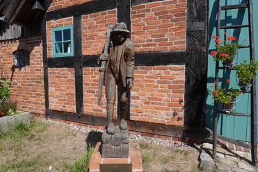 Skulptur "Der Häusling" Holzskulptur eines Mannes mit Stock vor einer Backsteinmauer, flankiert von Fenster und Blumenkasten.
