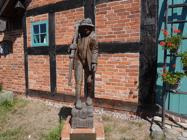 Skulptur "Der Häusling" Holzskulptur eines Mannes mit Stock vor einer Backsteinmauer, flankiert von Fenster und Blumenkasten.