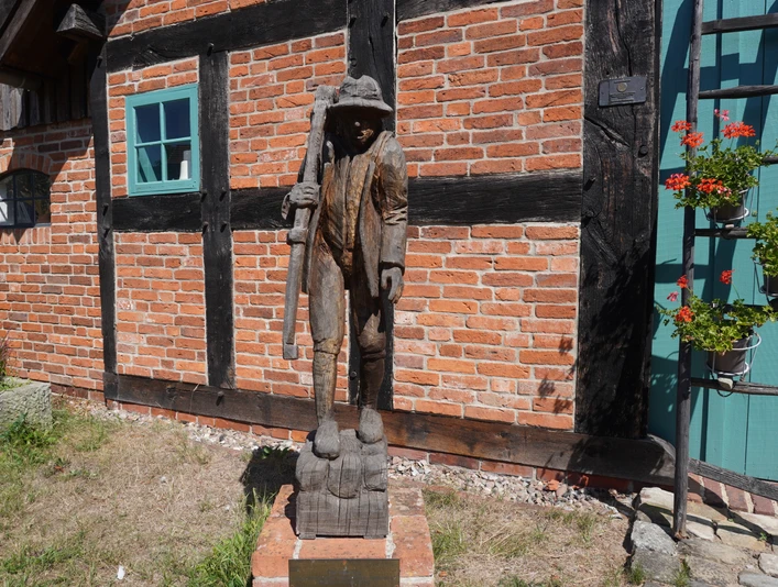 Skulptur "Der Häusling" Holzskulptur eines Mannes mit Stock vor einer Backsteinmauer, flankiert von Fenster und Blumenkasten.