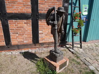 Skulptur "Der Häusling" Eine hölzerne Skulptur eines Mannes mit Stock vor einem Fachwerkgebäude, daneben blühende Pflanzen.