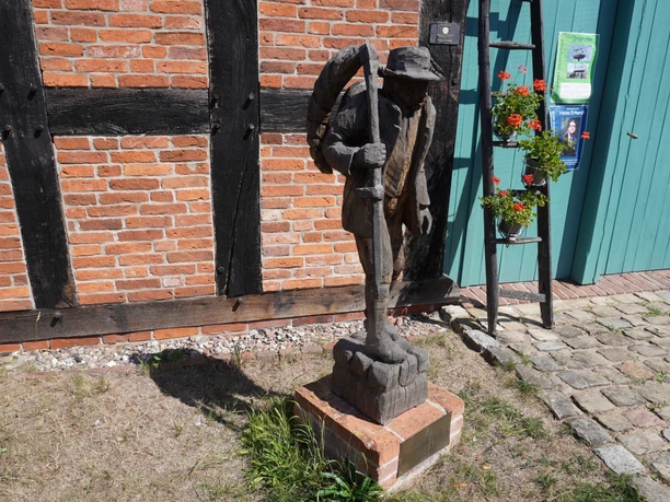 Skulptur "Der Häusling" Eine hölzerne Skulptur eines Mannes mit Stock vor einem Fachwerkgebäude, daneben blühende Pflanzen.