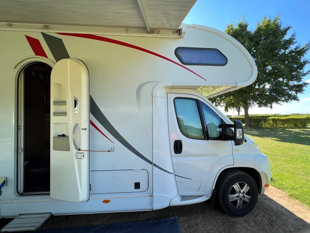 WoMo Ein Wohnmobil steht auf einem sonnigen Campingplatz mit geöffnetem Einstieg; im Hintergrund Bäume.
