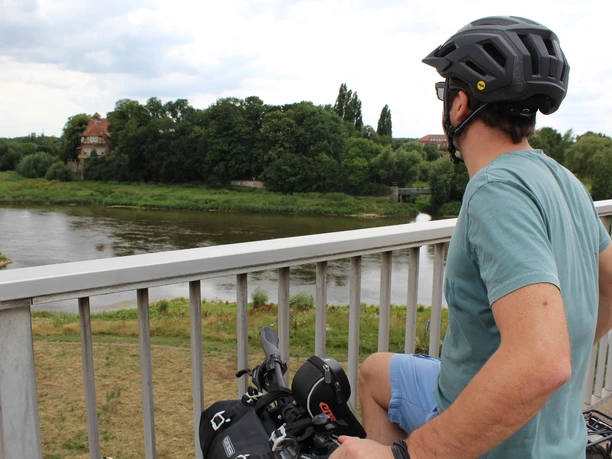 Radfahrer blickt von einer Brücke über die Weser auf üppige grüne Landschaft und ferne Gebäude.