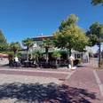 Brasserie am Markt in Apen Die Brasserie am Markt in Apen präsentiert sich mit einer einladenden Terrasse, gesäumt von Palmen, die unter strahlend blauem Himmel Schatten spenden. Vorne auf dem Platz fährt ein Radfahrer vorbei, während das Gebäude im Hintergrund eine lebendige kulinarische Atmosphäre verspricht.