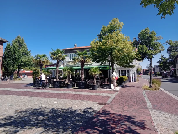 Brasserie am Markt in Apen Die Brasserie am Markt in Apen präsentiert sich mit einer einladenden Terrasse, gesäumt von Palmen, die unter strahlend blauem Himmel Schatten spenden. Vorne auf dem Platz fährt ein Radfahrer vorbei, während das Gebäude im Hintergrund eine lebendige kulinarische Atmosphäre verspricht.