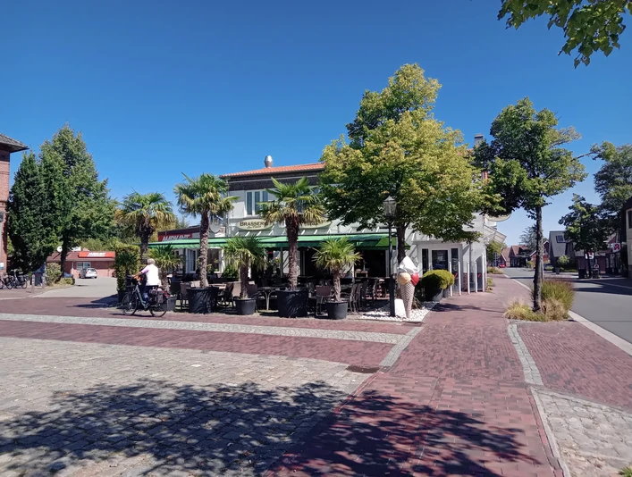 Brasserie am Markt in Apen Die Brasserie am Markt in Apen präsentiert sich mit einer einladenden Terrasse, gesäumt von Palmen, die unter strahlend blauem Himmel Schatten spenden. Vorne auf dem Platz fährt ein Radfahrer vorbei, während das Gebäude im Hintergrund eine lebendige kulinarische Atmosphäre verspricht.