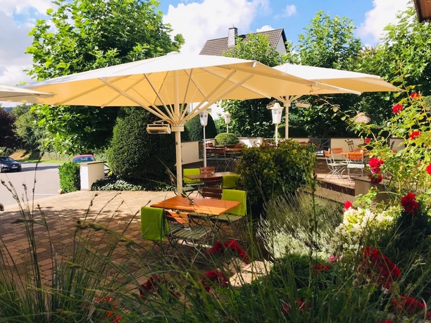 Terrasse Landhotel Weserblick Gepflasterte Terrasse mit Biergartenstühlen, grünen Decken und Sitzkissen, Tische aus Holz, weiße Sonnenschirme zur Beschattung, Tische umgeben von Grün-und Blühpflanzen