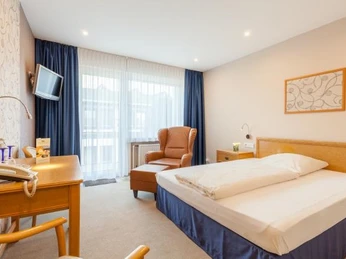 Zimmer mit Doppelbett, Kopfende aus Holz, beigefarbener Ohrensessel mit Fußbank, weiße Garden und blaue Übergardinen, links steht ein Schreibtisch und Stuhl aus Holz, Fernseher mit Wandhalterung