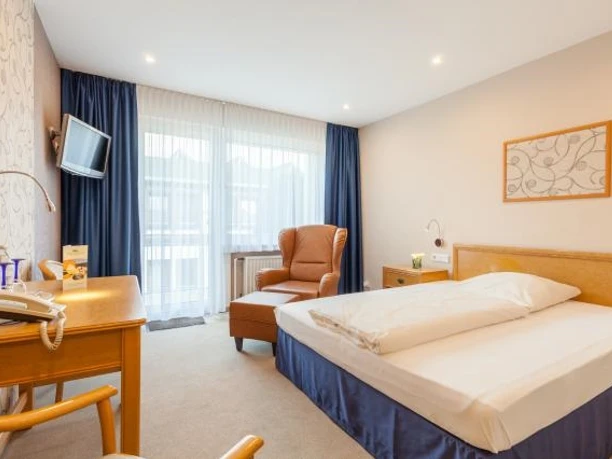 Zimmer mit Doppelbett, Kopfende aus Holz, beigefarbener Ohrensessel mit Fußbank, weiße Garden und blaue Übergardinen, links steht ein Schreibtisch und Stuhl aus Holz, Fernseher mit Wandhalterung