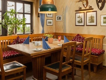 Gedeckter Holztisch mit drei Holzstühlen, umlaufende Sitzbank aus Holz mit floraler Polsterauflage, florale Kissen in grau-rot, Fotos in braunen Rahmen an der Wand, Fenster im Hintergrund