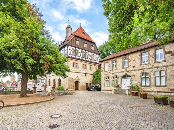 Rathaus zwischen den Städten (Ruben Emme) Fachwerkgebäude mit Uhrenturm, Kopfsteinpflaster und grünen Bäumen unter blauem Himmel in einer Stadt.
