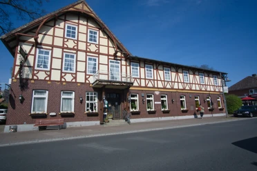 Außen.jpg Traditionelles Fachwerkhaus mit Backstein-Erdgeschoss an einer Straße unter blauem Himmel.
