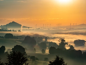 Nebeldurchzogene Hügellandschaft bei Sonnenaufgang, Windräder am Horizont, Desenberg im Hintergrund.