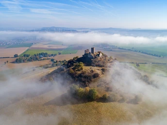 Ruine Desenberg auf sanfter Hügelkuppe, umgeben von Landschaft und Nebelschwaden, bei blauem Himmel.