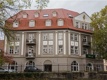 Historisches Gebäude mit rotem Ziegeldach, weißen Fensterrahmen und Balkonen, umgeben von Bäumen.