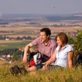 Ein Paar sitzt auf einer Wiese vor einer weiten Landschaft, entspannt, mit Blick auf Felder.