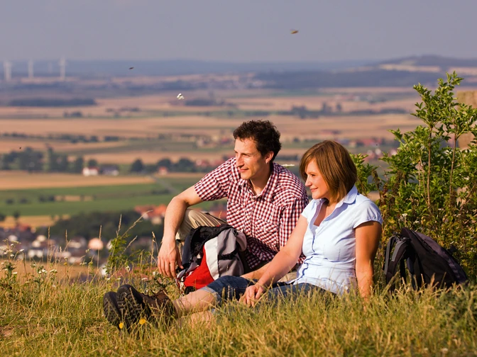 Ein Paar sitzt auf einer Wiese vor einer weiten Landschaft, entspannt, mit Blick auf Felder.