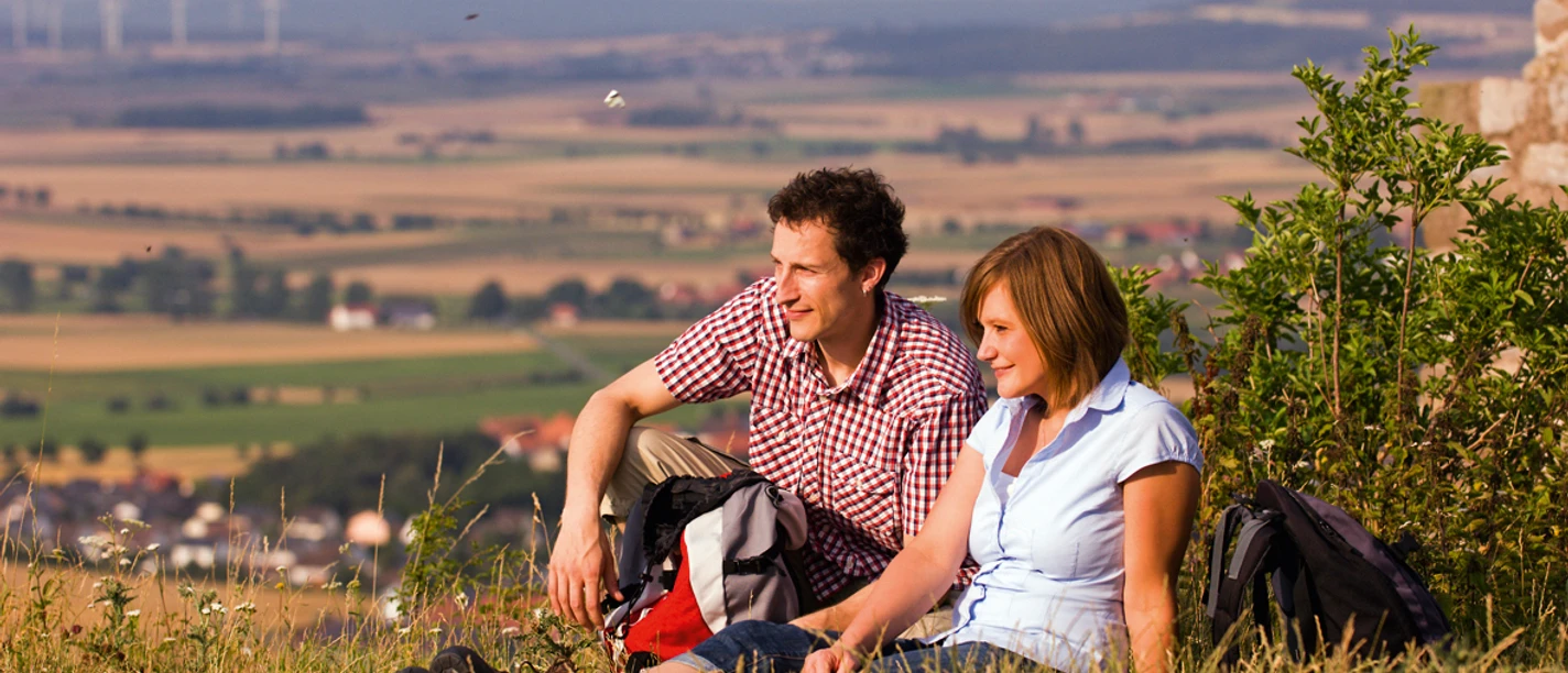 Ein Paar sitzt auf einer Wiese vor einer weiten Landschaft, entspannt, mit Blick auf Felder.