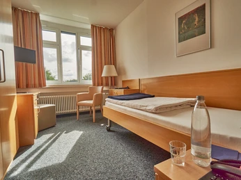 Klinik Berlin Zimmer.jpg Ein Einzelbettzimmer mit Holzmöbeln, großen Fenstern und einer hellen, freundlich gestalteten Atmosphäre.