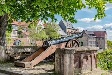 Fügeler-Kanone (Ruben Emme) Historische Kanone unter Bäumen mit Blick auf Fachwerkhäuser und eine alte Stadtmauer im Hintergrund.