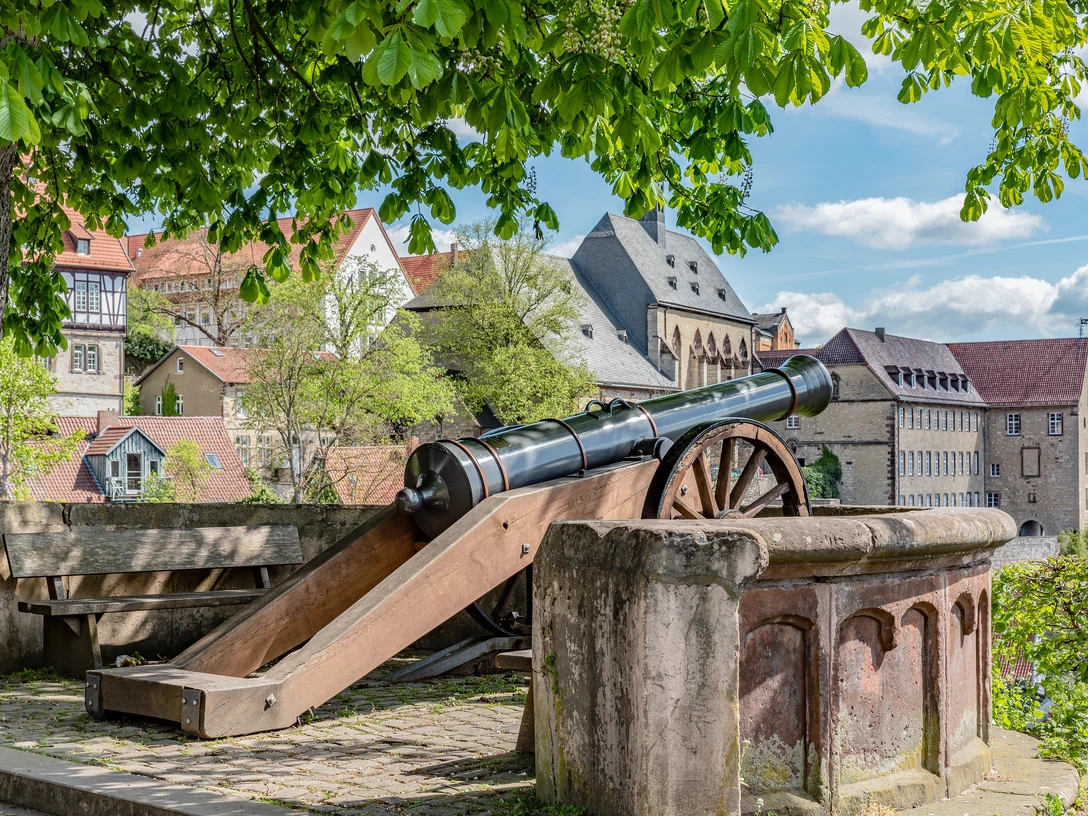 Fügeler-Kanone (Ruben Emme) Historische Kanone unter Bäumen mit Blick auf Fachwerkhäuser und eine alte Stadtmauer im Hintergrund.