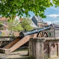 Fügeler-Kanone (Ruben Emme) Historische Kanone unter Bäumen mit Blick auf Fachwerkhäuser und eine alte Stadtmauer im Hintergrund.