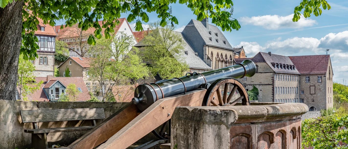 Fügeler-Kanone (Ruben Emme) Historische Kanone unter Bäumen mit Blick auf Fachwerkhäuser und eine alte Stadtmauer im Hintergrund.